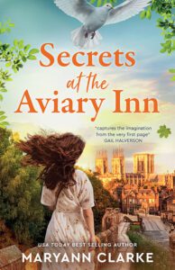 dark haired young woman in floral dress walking along city wall with York, England and Minster cathedral in the distance. A white dove hovers at top. Orange serif font title: Secrets at the Aviary Inn. USA Today Best Selling Author MaryAnn Clarke in white block font at the bottom